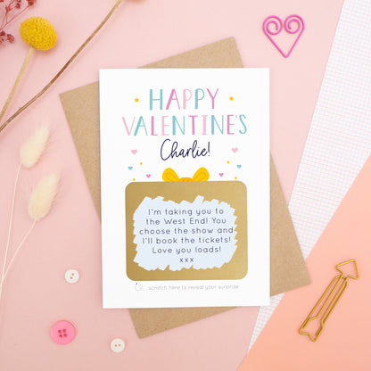 A personalised happy Valentine’s scratch card photographed on a pink background with floral props, paper clips, and buttons. The card has been scratched off to reveal the hidden message and is in the pink and blue colour scheme.