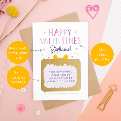 A personalised happy Valentine’s scratch card photographed on a pink background with floral props, paper clips, and buttons. The card has been scratched off to reveal the hidden message and is in the pink and lilac colour scheme. Around the card are circles of text pointing to the areas that can be changed (name, colour and personalised message).
