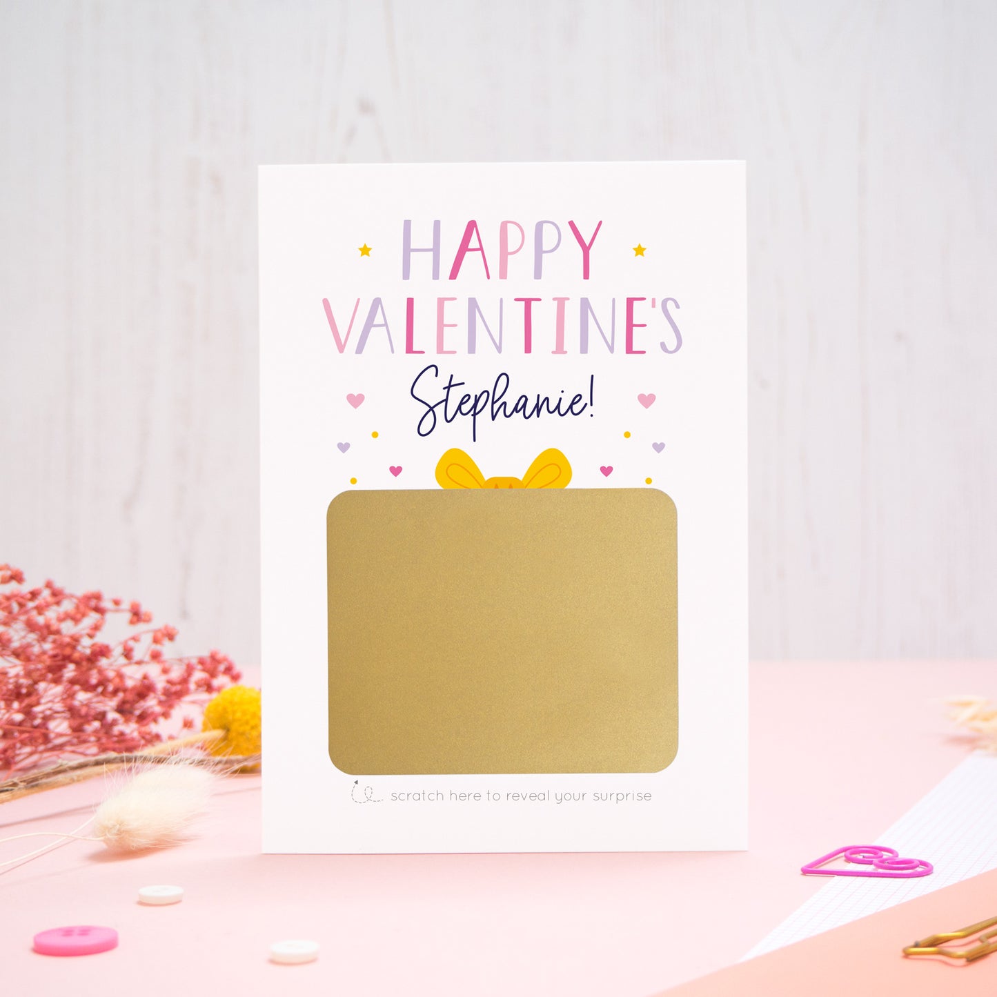 A personalised happy Valentine’s scratch card photographed on a pink background with floral props, paper clips, and buttons. The card has not yet been scratched off and is in the pink and lilac colour scheme.