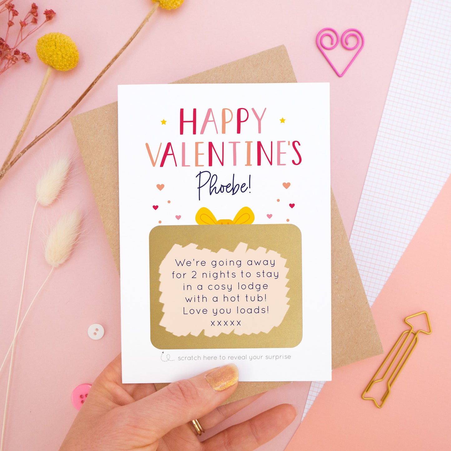 A personalised happy Valentine’s scratch card photographed on a pink background with floral props, paper clips, and buttons. The card has been scratched off to reveal the hidden message and is in the red and peach colour scheme.