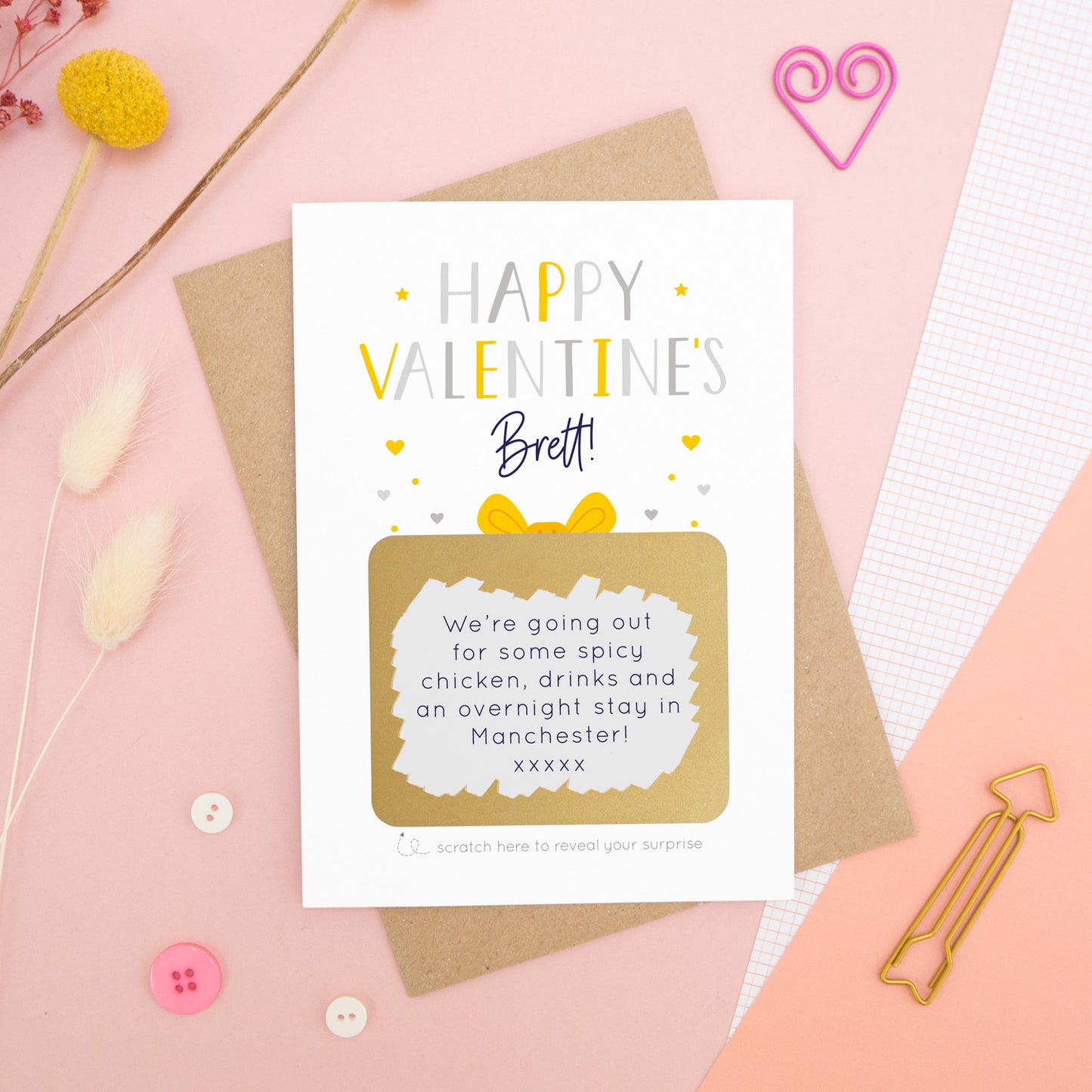 A personalised happy Valentine’s scratch card photographed on a pink background with floral props, paper clips, and buttons. The card has been scratched off to reveal the hidden message and is in the yellow and grey colour scheme.
