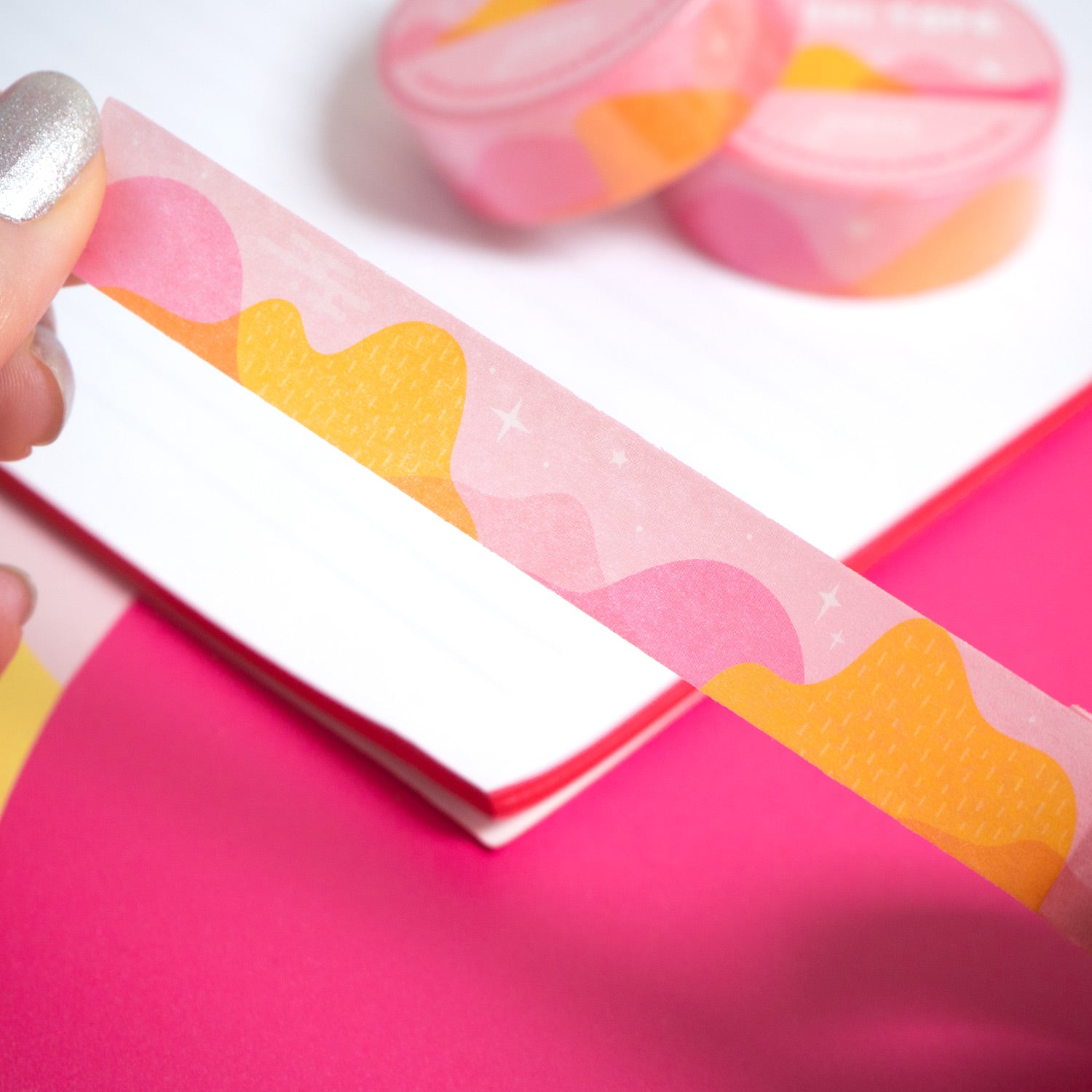 Desert mountain washi tape in pink and yellow being peeled from the roll by a pair of hands over a white notebook and pink background.