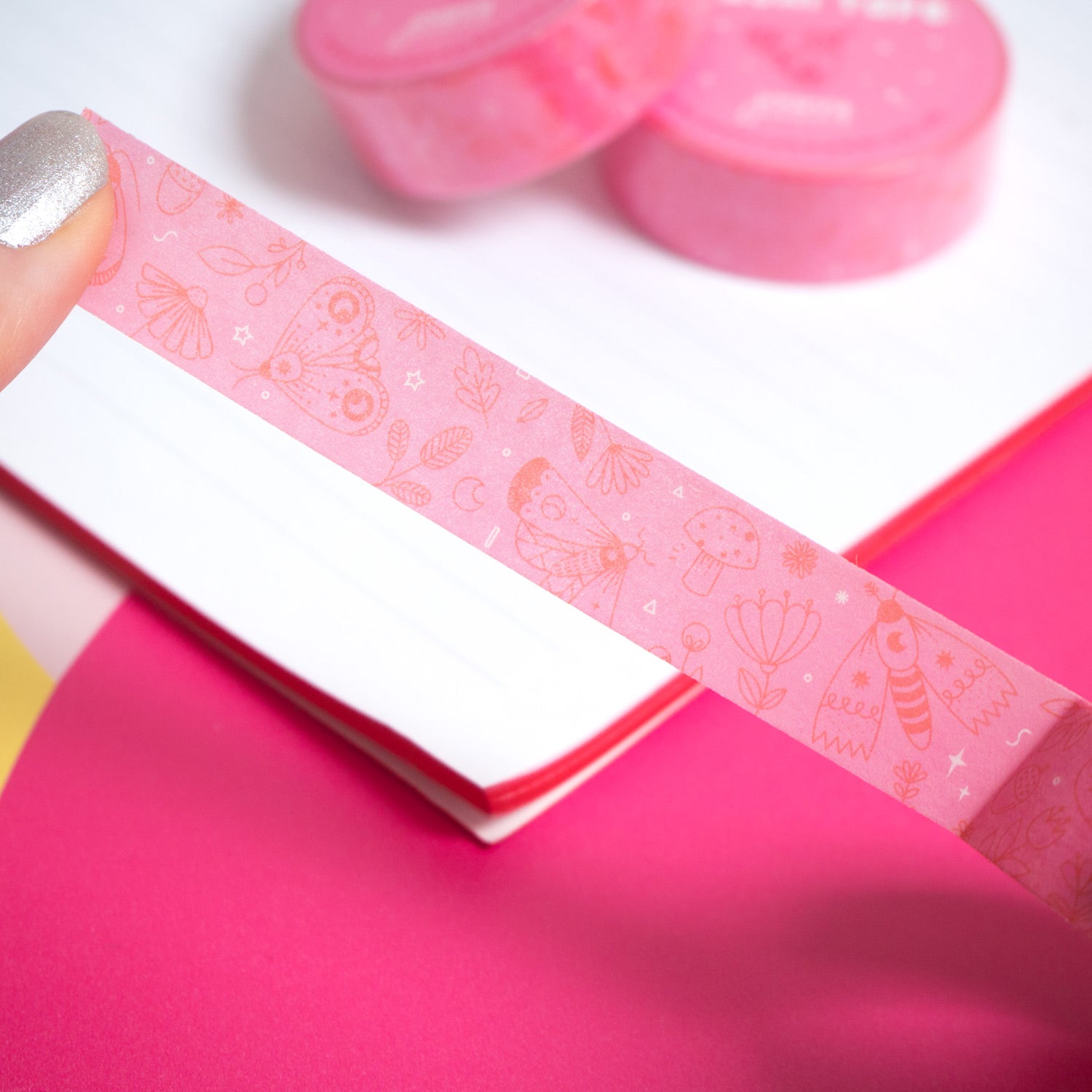 The pink moths washi tape being peeled from the roll by a pair of hands over a white notebook and pink background.