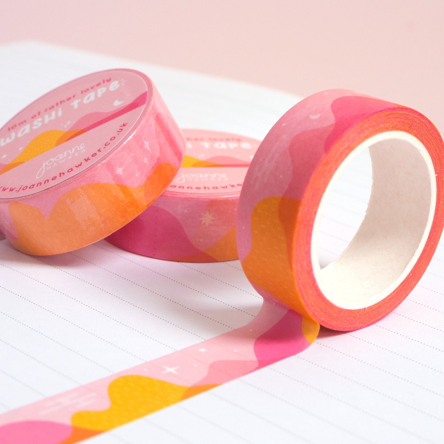 A roll of pink and yellow desert mountain washi tape unravelling on a notebook with stacks of paper tape in the background.