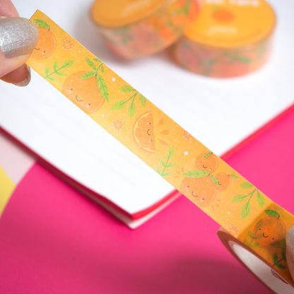 Cute orange washi tape being peeled from the roll by a pair of hands over a white notebook and pink background.