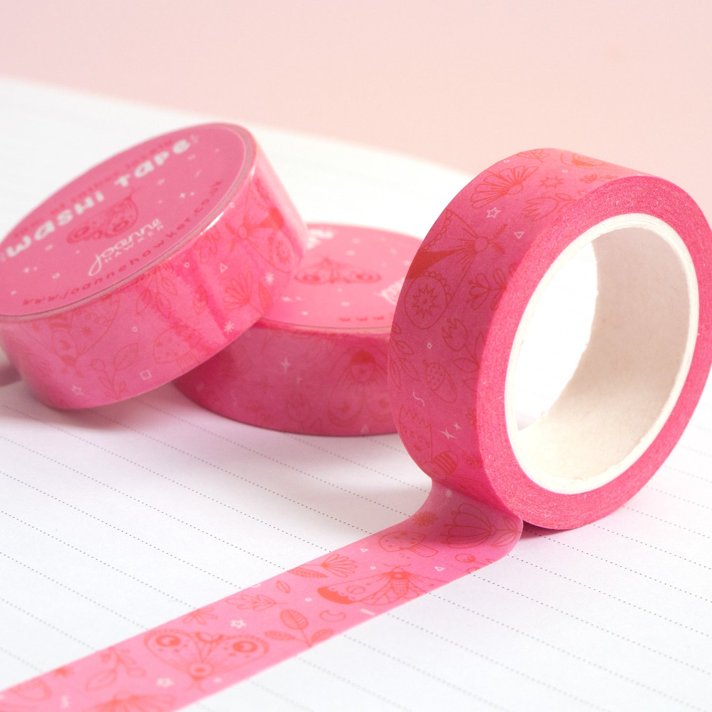 A roll of pink moths washi tape unravelling on a notebook with stacks of paper tape in the background.