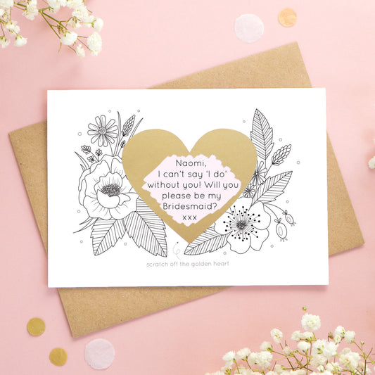 A personalised wedding scratch card shot on a pink background with white flowers. The golden heart has been scratched revealing a pink heart and a bridesmaid proposal!