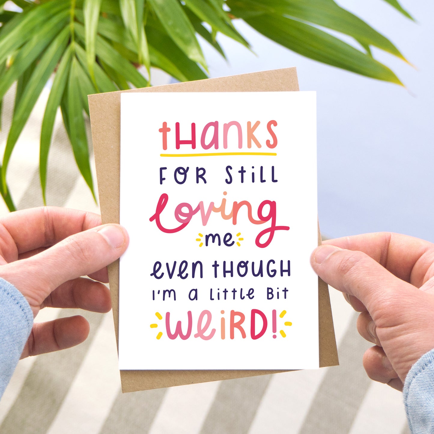 Thanks for still loving me even though I'm a little bit weird card in pink being held over a blue background with a leafy plant.