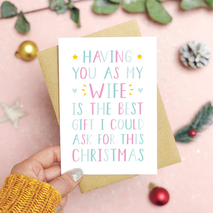 A 'best gift' wife Christmas card held over a pink background by a hand in a mustard knit jumper with foliage and baubles in the background. The writing on the card is blue and pink