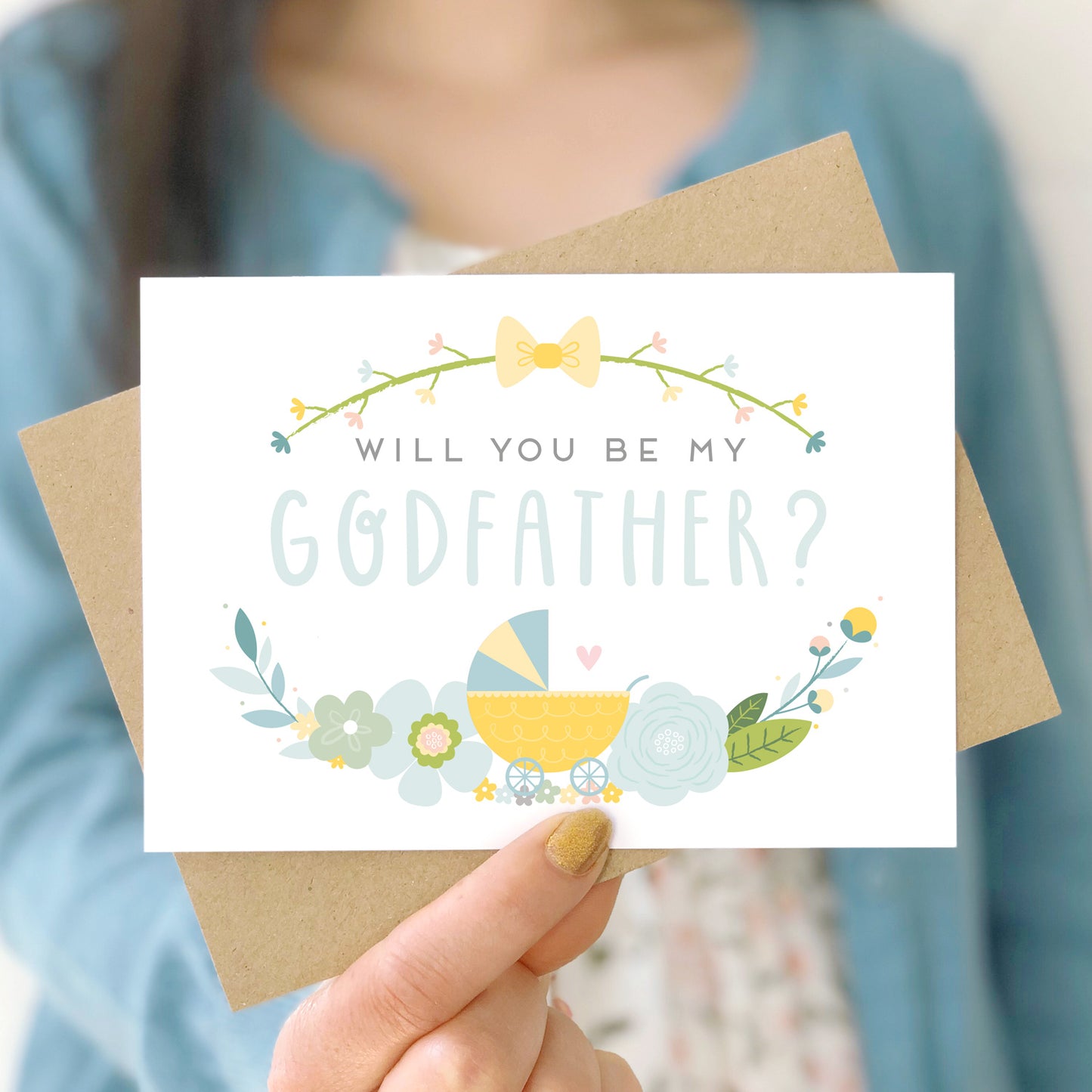 A will you be my godfather card being held in front of a white dress and blue cardigan. The design features a pram, simple florals and the all important question. This is the blue palette.