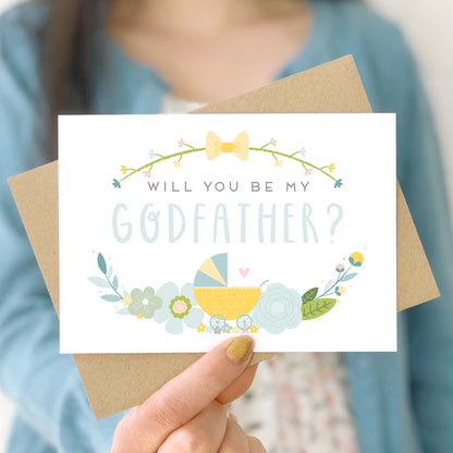 A will you be my godfather card being held in front of a white dress and blue cardigan. The design features a pram, simple florals and the all important question. This is the blue palette.