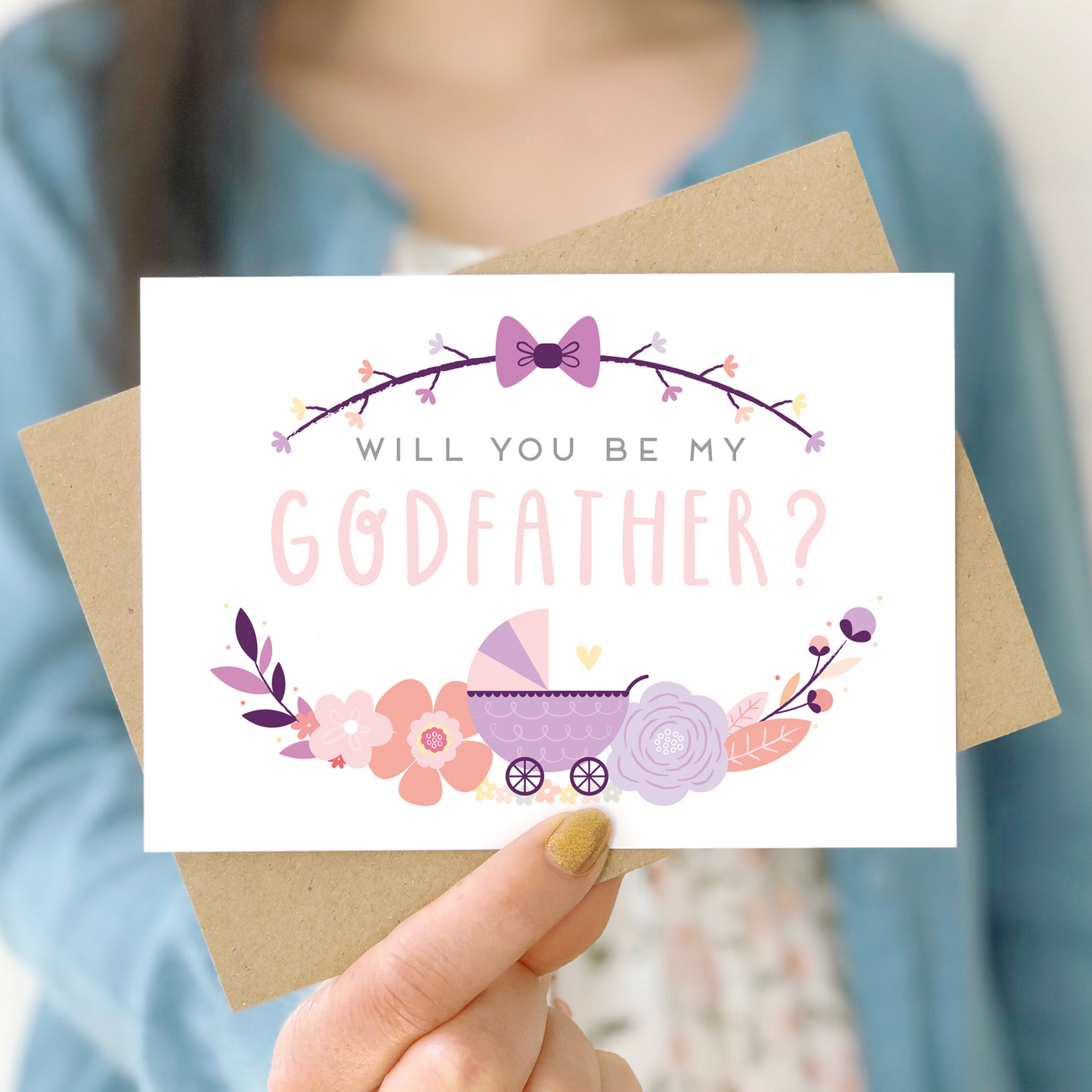 A will you be my godfather card being held in front of a white dress and blue cardigan. The design features a pram, simple florals and the all important question. This is the purple palette.