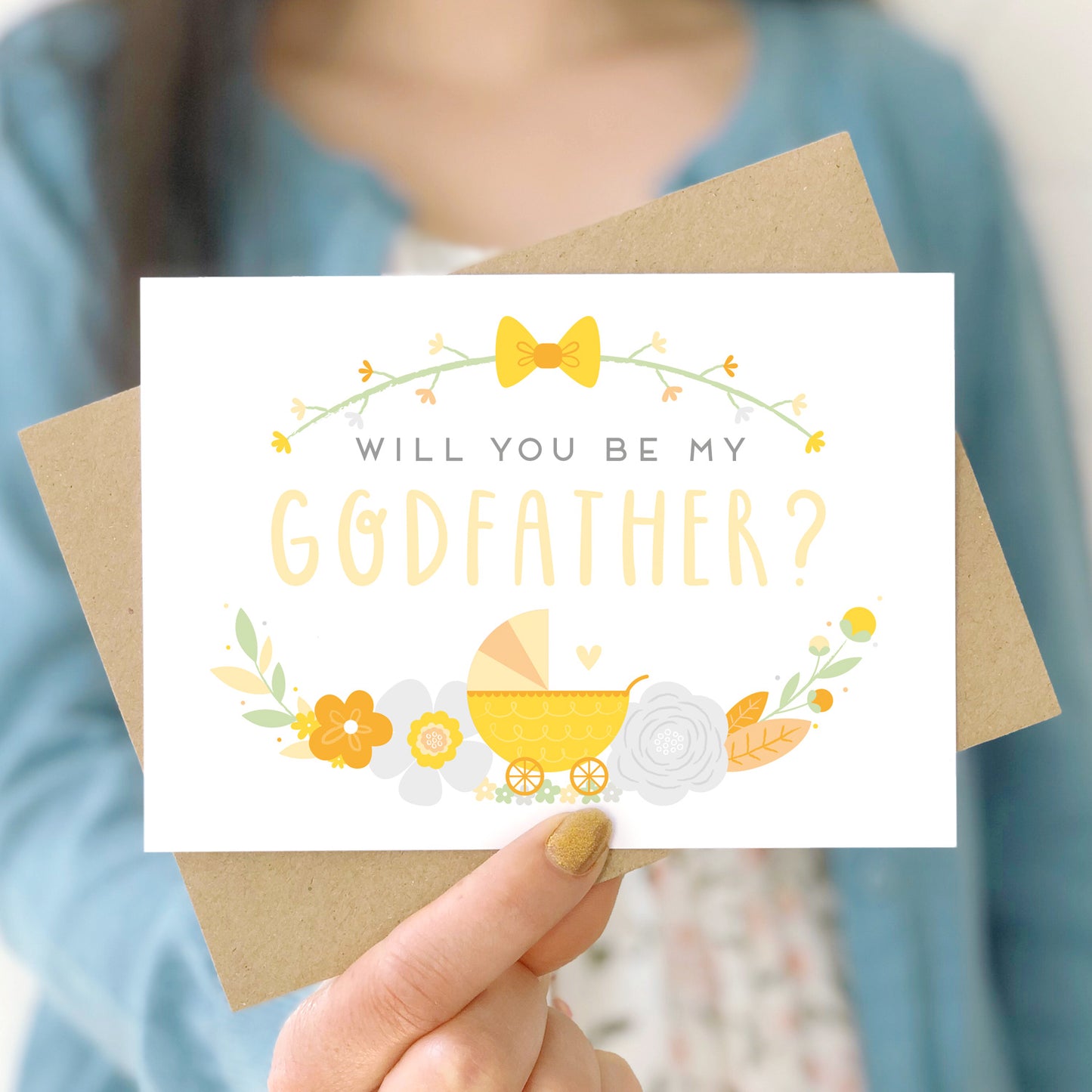 A will you be my godfather card being held in front of a white dress and blue cardigan. The design features a pram, simple florals and the all important question. This is the yellow palette.