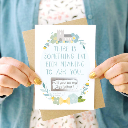 A will you be my godfather scratch and reveal card being held in front of a white dress and blue cardigan. The design features a church, simple florals and a scratch off panel in silver. This is the blue palette.