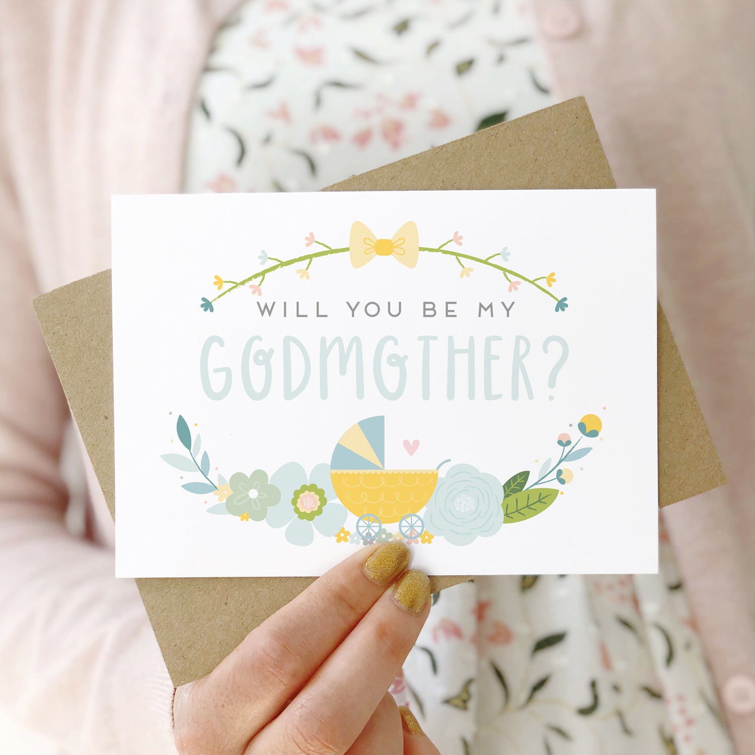 A will you be my godmother card being held in front of a white dress and pink cardigan. The design features a pram, simple florals and the all important question. This is the blue palette.
