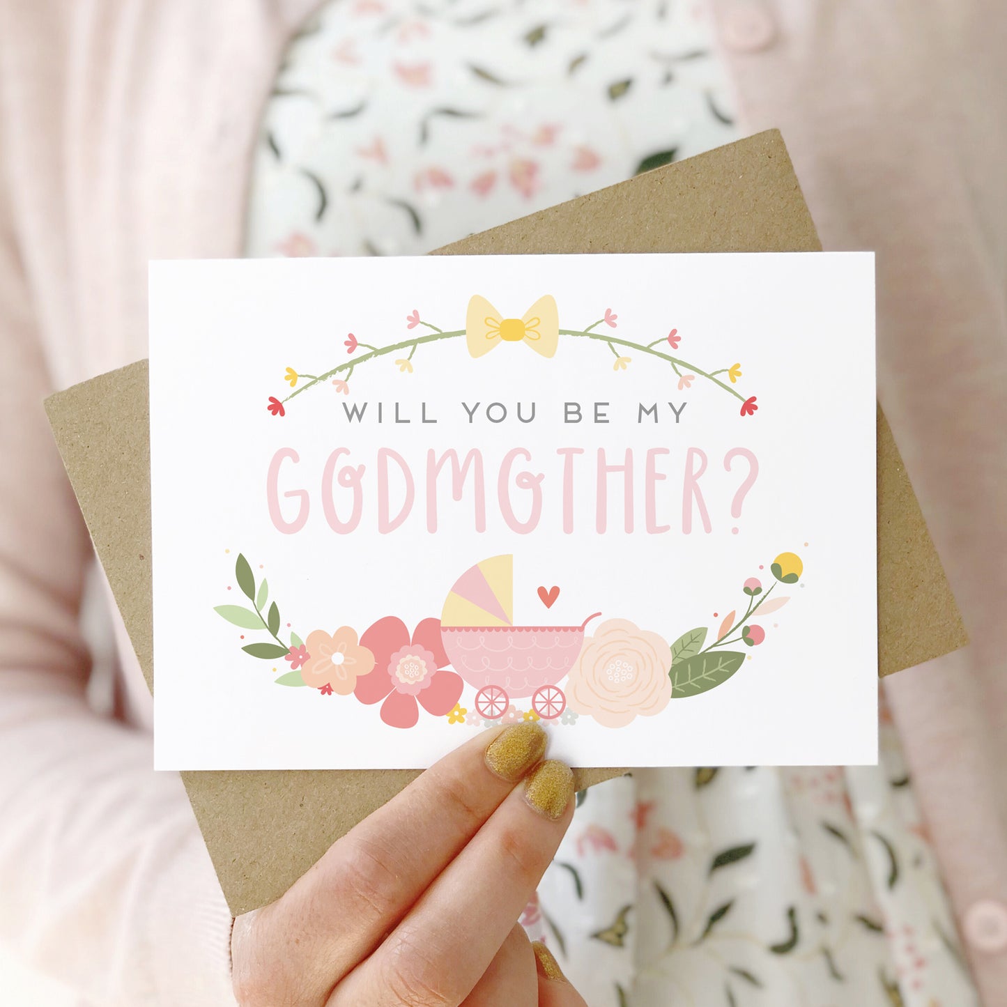 A will you be my godmother card being held in front of a white dress and pink cardigan. The design features a pram, simple florals and the all important question. This is the pink palette.