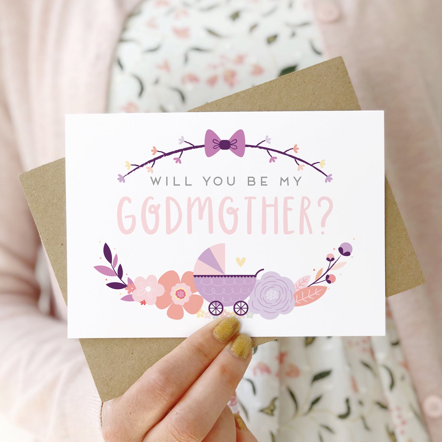 A will you be my godmother card being held in front of a white dress and pink cardigan. The design features a pram, simple florals and the all important question. This is the purple palette.