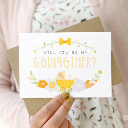 A will you be my godmother card being held in front of a white dress and pink cardigan. The design features a pram, simple florals and the all important question. This is the yellow palette.