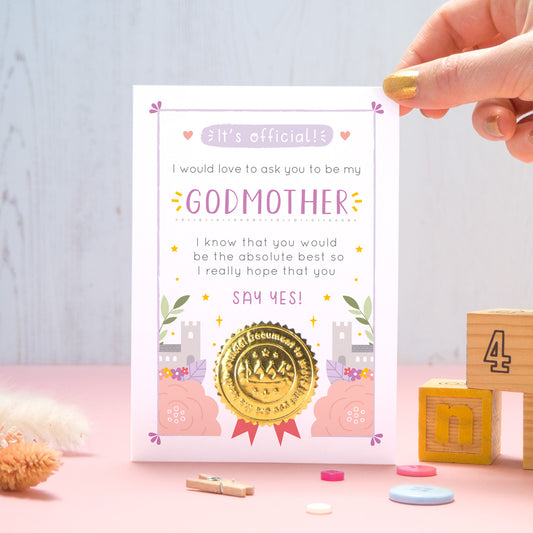 A will you be my Godmother certificate card in pink and purple, shot on a pink and grey background with a hand coming in from the top right. There are dry flowers, buttons and building blocks in the foreground.