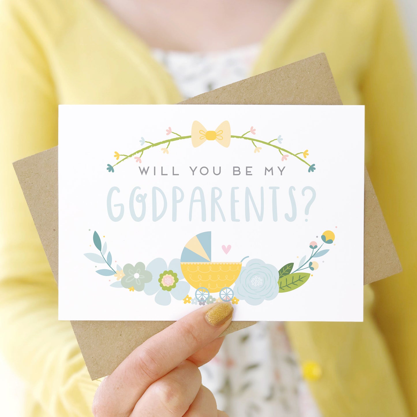 A will you be my godparents card being held in front of a white dress and yellow cardigan. The design features a pram, simple florals and the all important question. This is the blue palette.