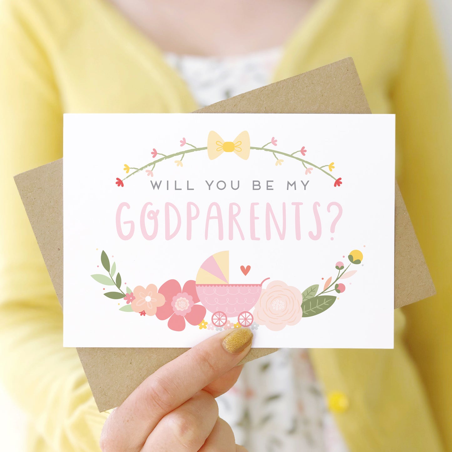 A will you be my godparents card being held in front of a white dress and yellow cardigan. The design features a pram, simple florals and the all important question. This is the pink palette.