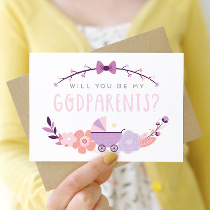 A will you be my godparents card being held in front of a white dress and yellow cardigan. The design features a pram, simple florals and the all important question. This is the purple palette.