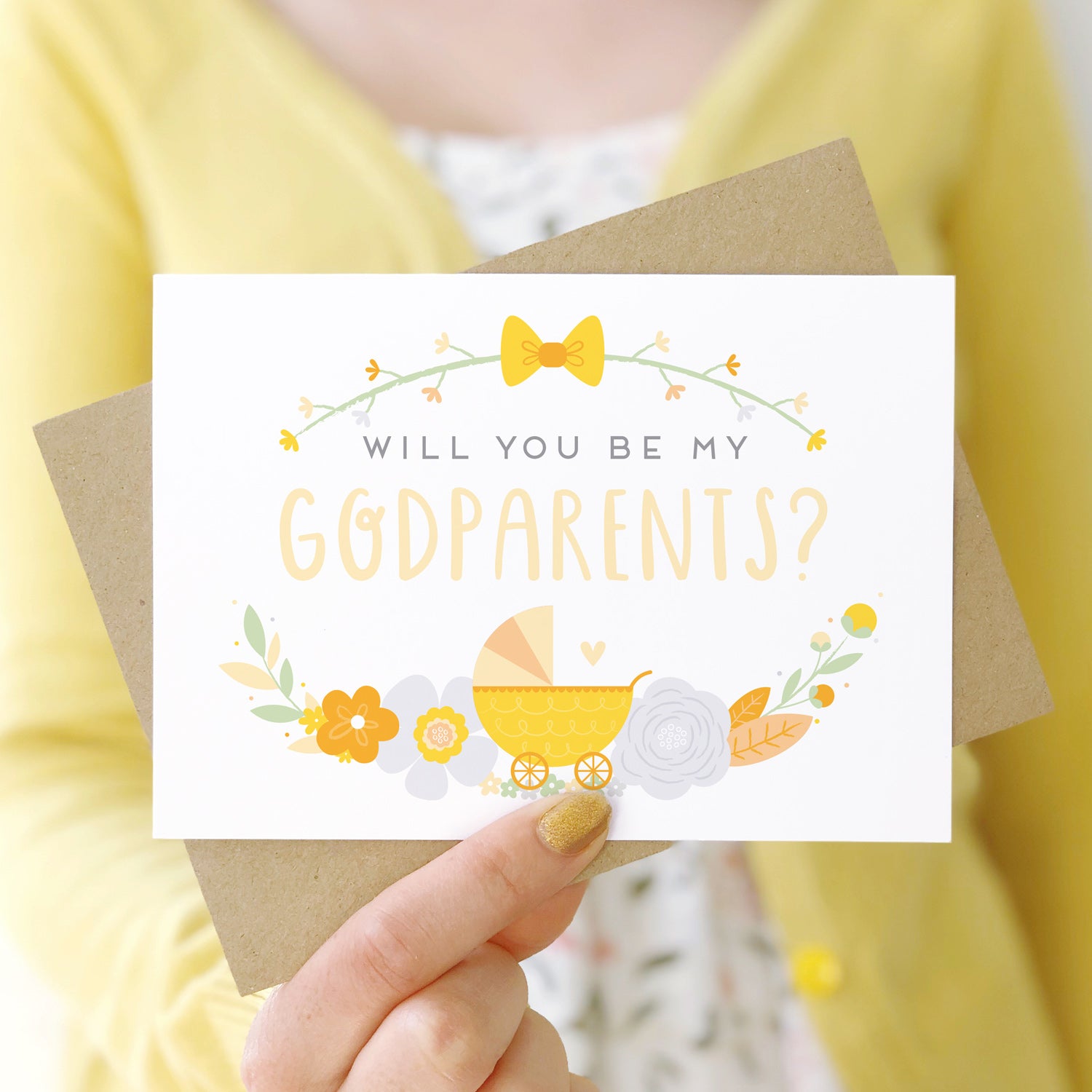 A will you be my godparents card being held in front of a white dress and yellow cardigan. The design features a pram, simple florals and the all important question. This is the yellow palette.