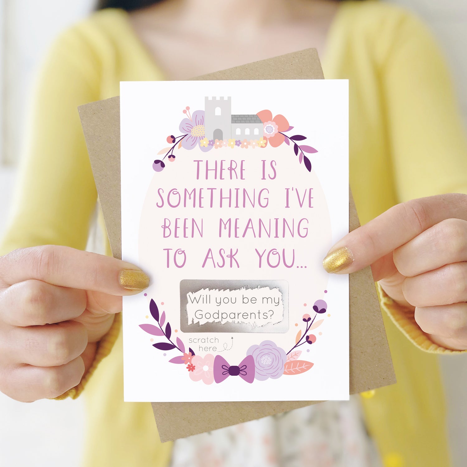 A will you be my godparents scratch and reveal card being held in front of a white dress and yellow cardigan. The design features a church, simple florals and a scratch off panel in silver. This is the purple palette.