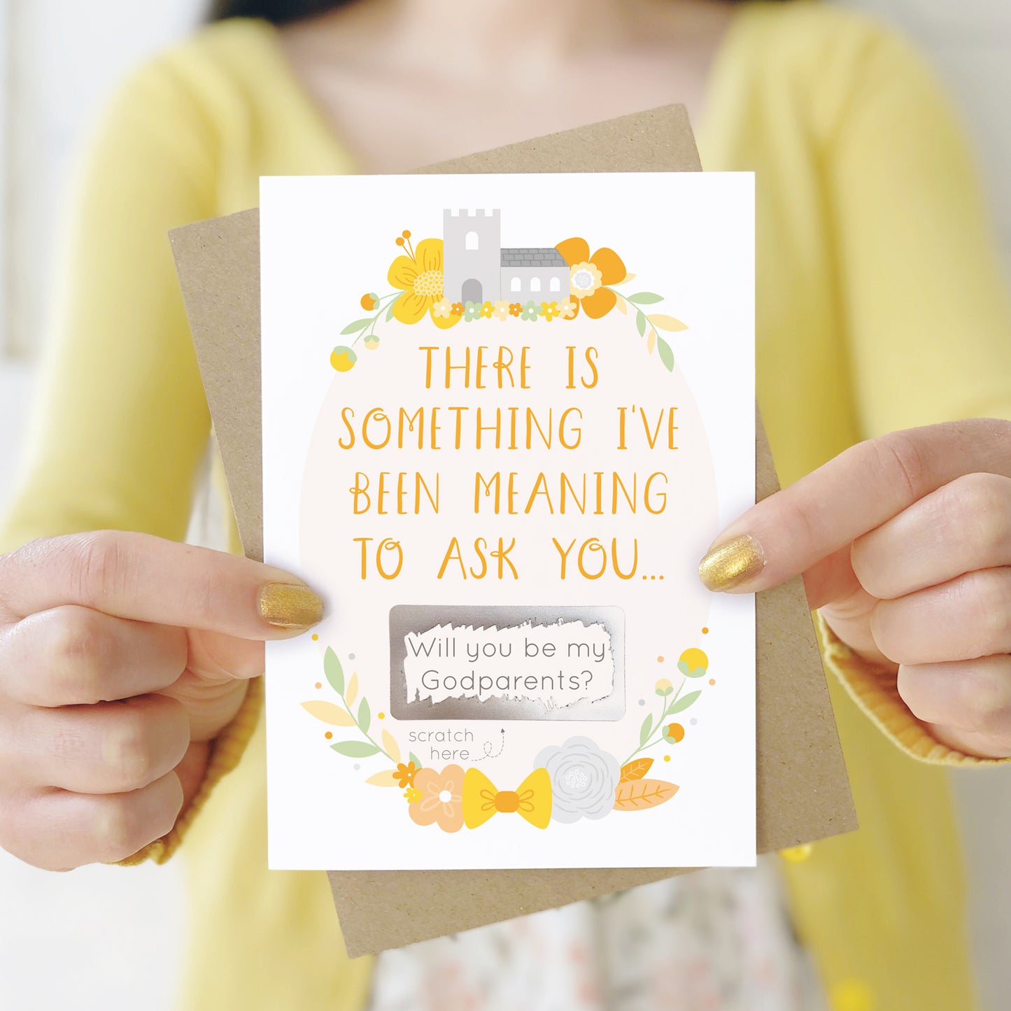 A will you be my godparents scratch and reveal card being held in front of a white dress and yellow cardigan. The design features a church, simple florals and a scratch off panel in silver. This is the yellow palette.