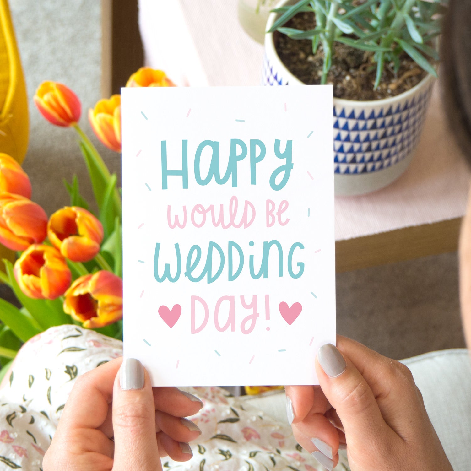 Happy would be wedding day card in teal and pink. Photographed in a lifestyle setting being held over a lap with tulips and a succulent in the background.