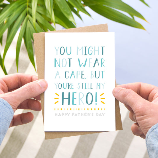 A hero father's day card in tones of blue and a pop of yellow photographed being held by a man over a grey stripy rug, blue floor and a leafy plant.