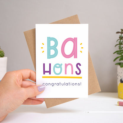 A Ba Hons graduation congratulations card held over a warm grey and white background with potted plants peeping the sides. Behind the card is a kraft brown envelope that comes with the card. The text on this version of the card is in varying tones of navy and pink, purple and  blue.