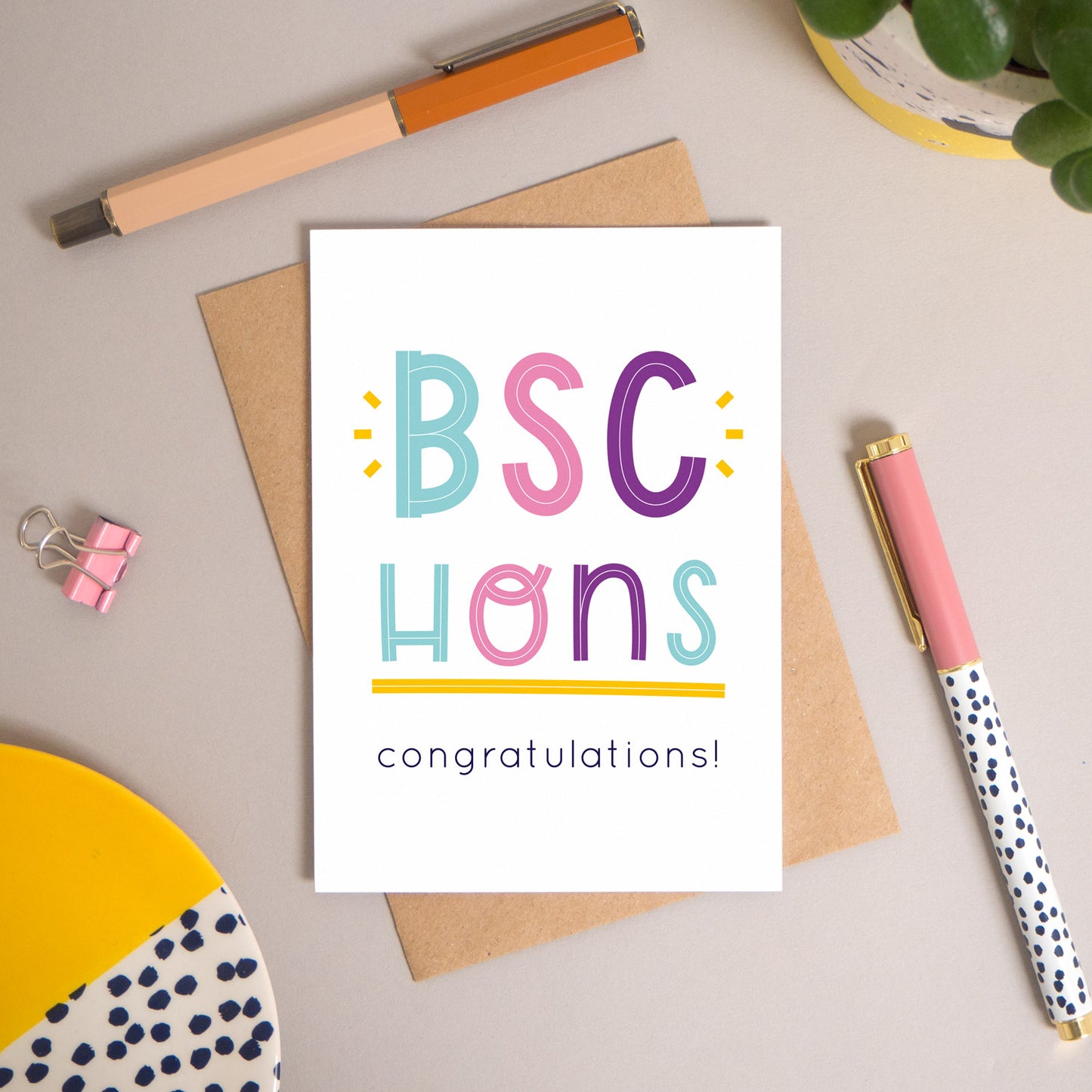 This BSc Hons graduation congratulations card has been shot flatlay style looking directly over the top of the card. It is lying flat on it’s kraft brown envelope, on a warm grey background. Surrounding the card are pens, a clip, a plant and a trinket dish. This version of the card is in varying tones of navy and pink, purple and blue.