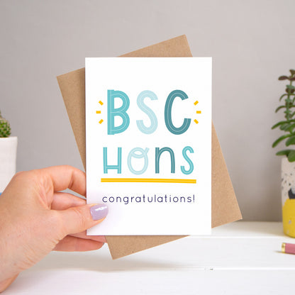 A BSc Hons graduation congratulations card held over a warm grey and white background with potted plants peeping the sides. Behind the card is a kraft brown envelope that comes with the card. The text on this version of the card is in varying tones of navy and blue.