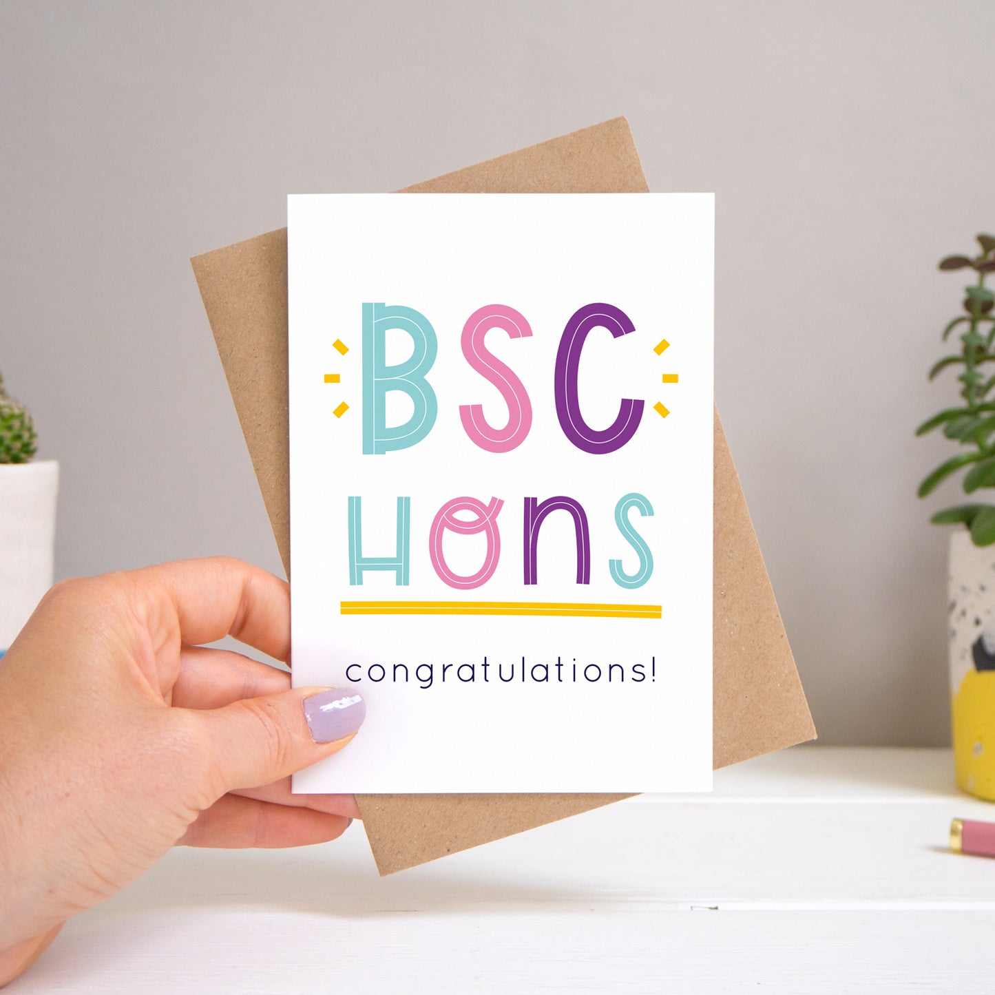 A BSc Hons graduation congratulations card held over a warm grey and white background with potted plants peeping the sides. Behind the card is a kraft brown envelope that comes with the card. The text on this version of the card is in varying tones of navy and pink, purple and  blue.