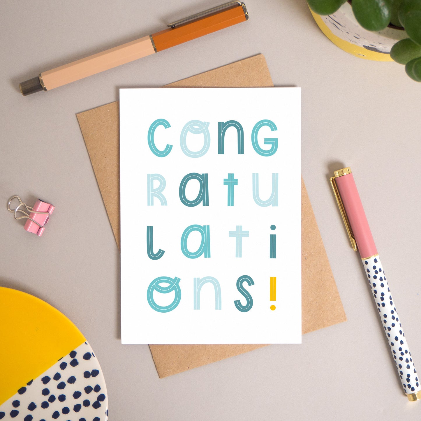 This congratulations card has been shot flatlay style looking directly over the top of the card. It is lying flat on it’s kraft brown envelope, on a warm grey background. Surrounding the card are pens, a clip, a plant and a trinket dish. This version of the card is in varying tones of blue.