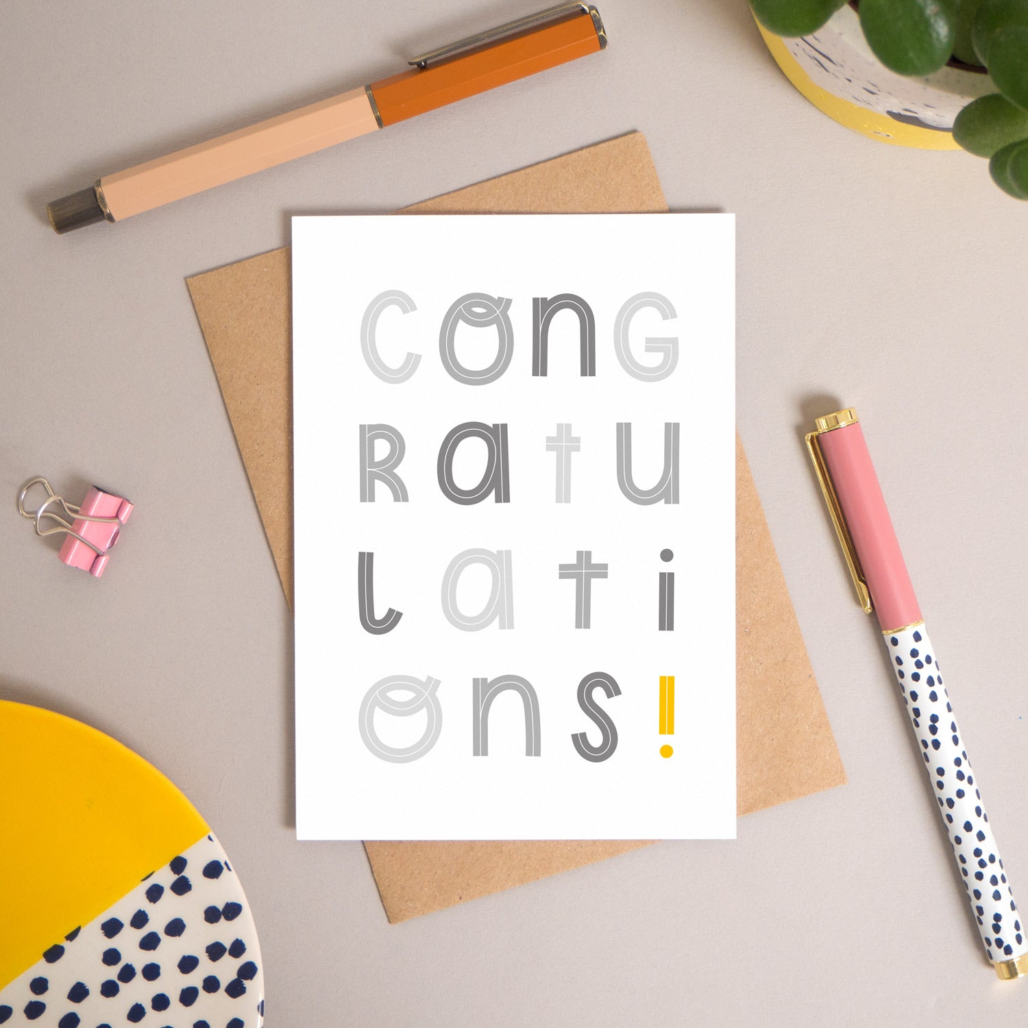 This congratulations card has been shot flatlay style looking directly over the top of the card. It is lying flat on it’s kraft brown envelope, on a warm grey background. Surrounding the card are pens, a clip, a plant and a trinket dish. This version of the card is in varying tones of grey.