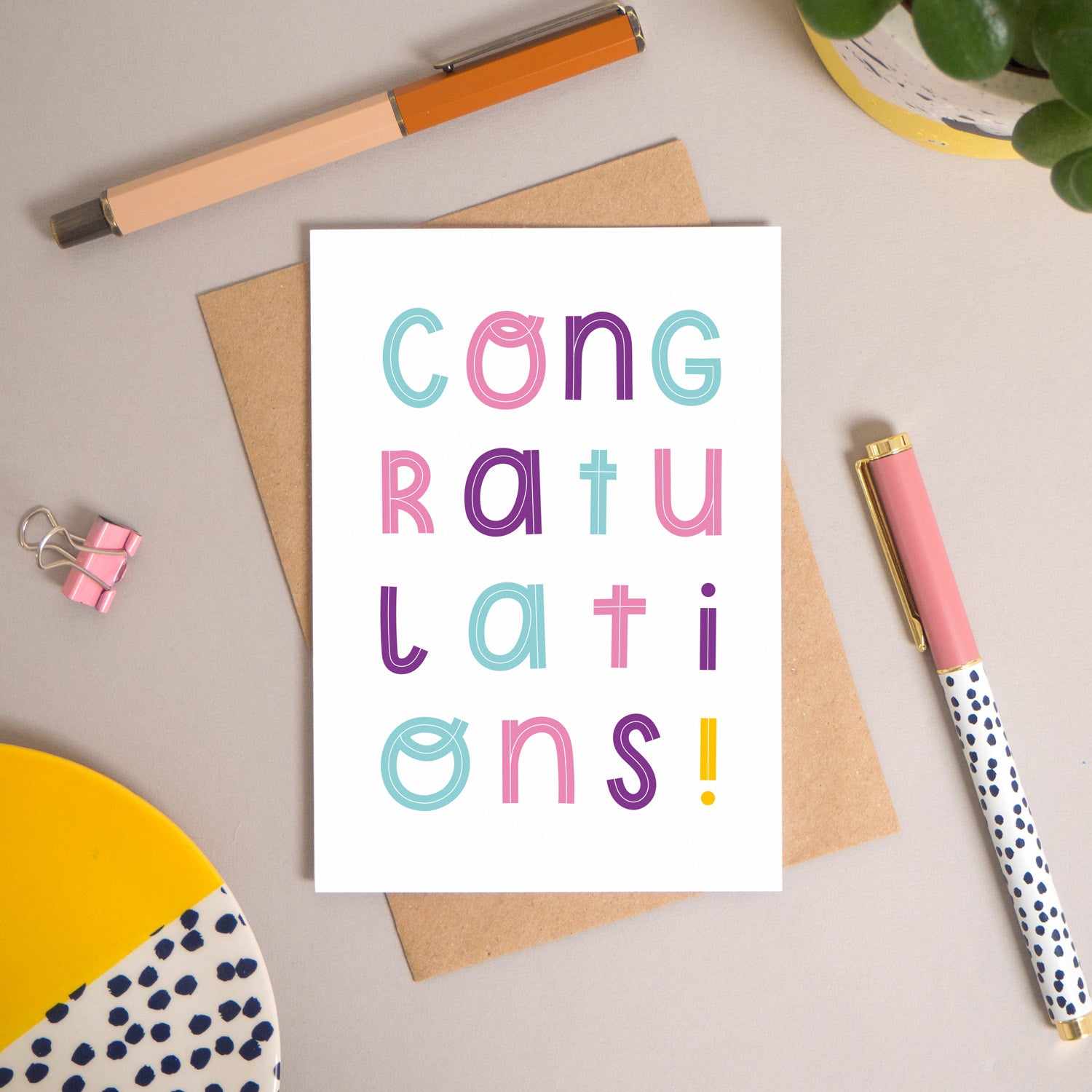 This congratulations card has been shot flatlay style looking directly over the top of the card. It is lying flat on it’s kraft brown envelope, on a warm grey background. Surrounding the card are pens, a clip, a plant and a trinket dish. This version of the card is in varying tones of pink, purple and blue.