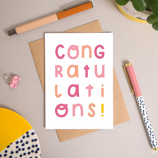This congratulations card has been shot flatlay style looking directly over the top of the card. It is lying flat on it’s kraft brown envelope, on a warm grey background. Surrounding the card are pens, a clip, a plant and a trinket dish. This version of the card is in varying tones of pink.