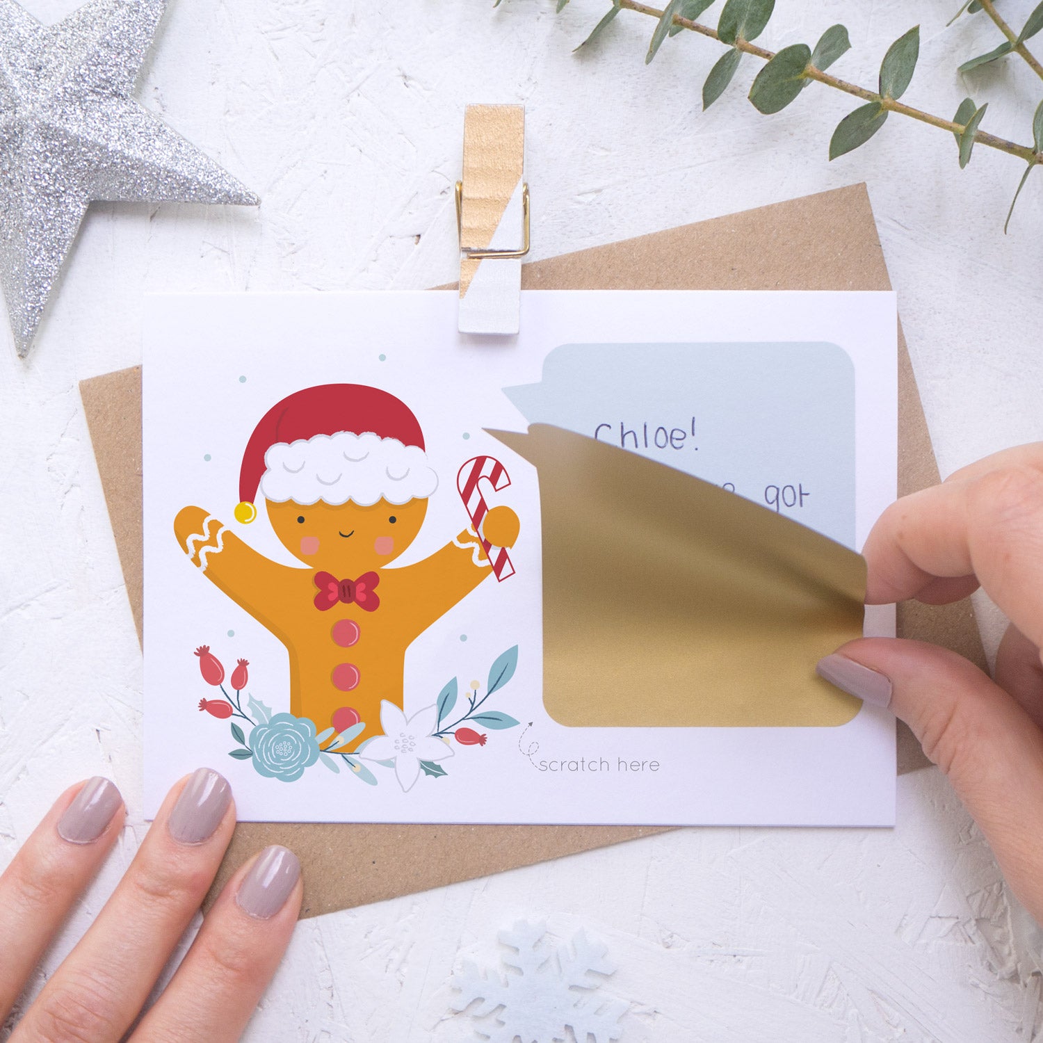 A personalised gingerbread man scratch card where the sticking down of the gold scratch panel is being demonstrated. Shot on a white background with a glittery star and sprig of eucalyptus.