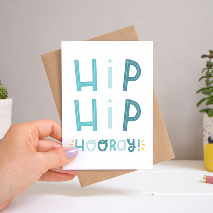 A ‘hip hip hooray!’ card held over a warm grey and white background with potted plants peeping the sides. Behind the card is a kraft brown envelope that comes with the card. The text on this version of the card is in varying tones of blue.