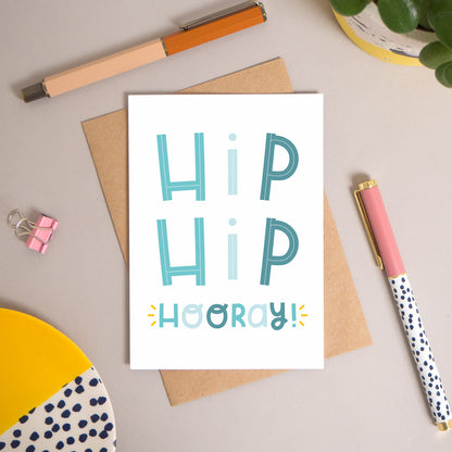 This ‘hip hip hooray!’ card has been shot flatlay style looking directly over the top of the card. It is lying flat on it’s kraft brown envelope, on a warm grey background. Surrounding the card are pens, a clip, a plant and a trinket dish. This version of the card is in varying tones of blue.