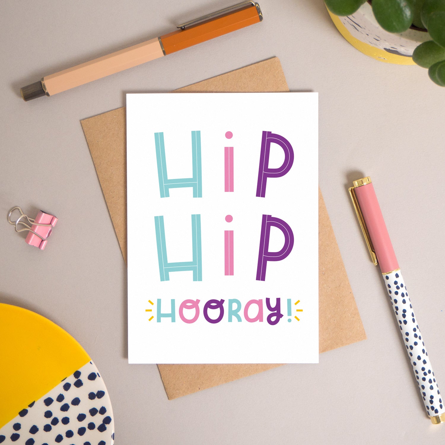 This ‘hip hip hooray!’ card has been shot flatlay style looking directly over the top of the card. It is lying flat on it’s kraft brown envelope, on a warm grey background. Surrounding the card are pens, a clip, a plant and a trinket dish. This version of the card is in varying tones of pink, purple and  blue.