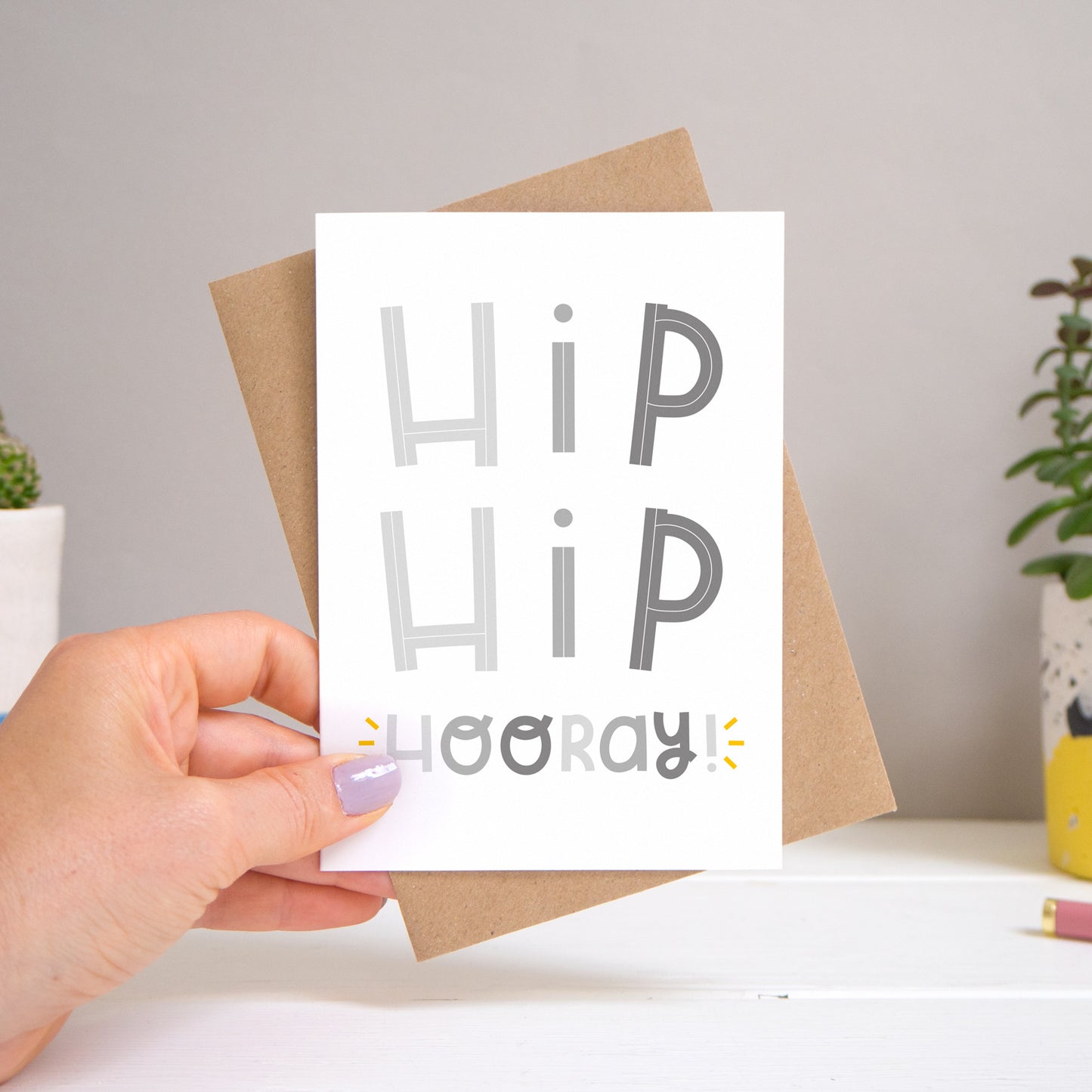 A ‘hip hip hooray!’ card held over a warm grey and white background with potted plants peeping the sides. Behind the card is a kraft brown envelope that comes with the card. The text on this version of the card is in varying tones of grey.