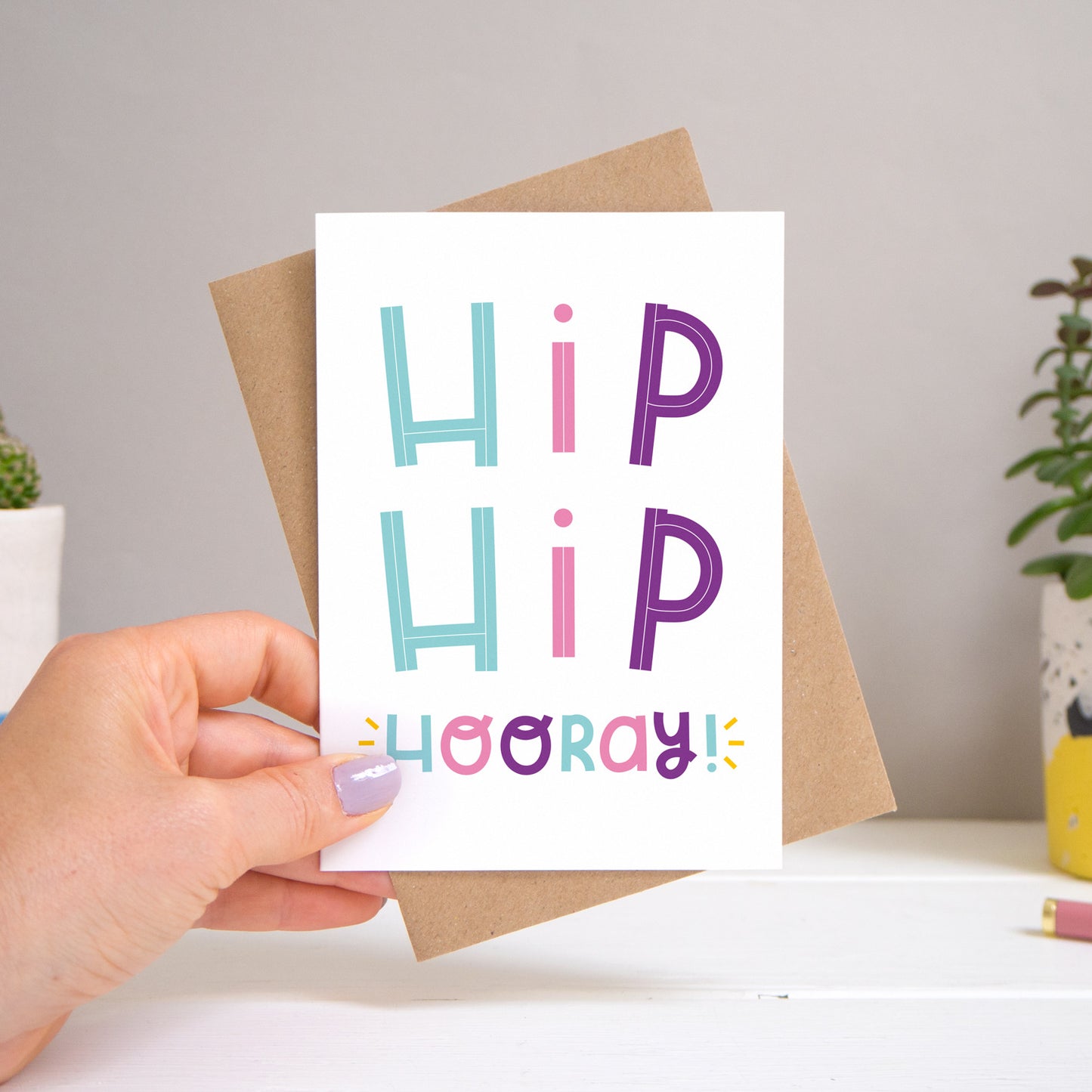 A ‘hip hip hooray!’ card held over a warm grey and white background with potted plants peeping the sides. Behind the card is a kraft brown envelope that comes with the card. The text on this version of the card is in varying tones of pink, purple and  blue.