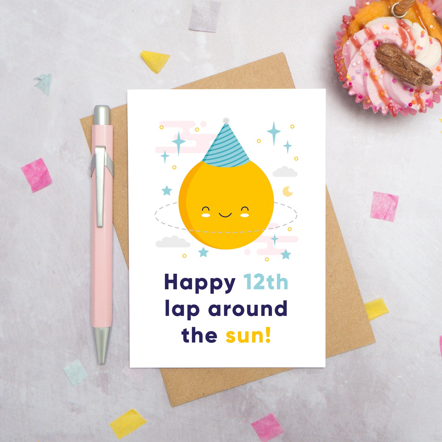 An any age birthday card featuring a happy sun in a party hat and text that reads 'Happy 12th lap around the sun'. This card has been photographed on a grey background surrounded by a cupcake, pen and confetti.