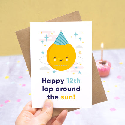 An any age birthday card featuring a happy sun in a party hat and text that reads 'Happy 12th lap around the sun'. This card has been photographed being held on a grey and yellow background with cupcake and candle in the distance.
