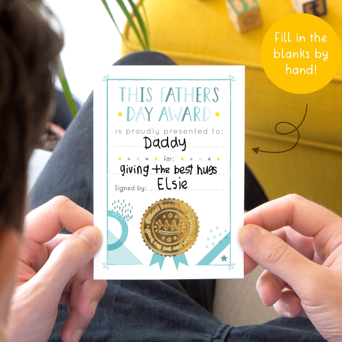 The Fathers day award card being held by a male on a sofa. There is a yellow footstool in the top right and he is holding the version of the card that a child has filled out for him. This card is blue and white in colour with a shiny gold certificate seal.
