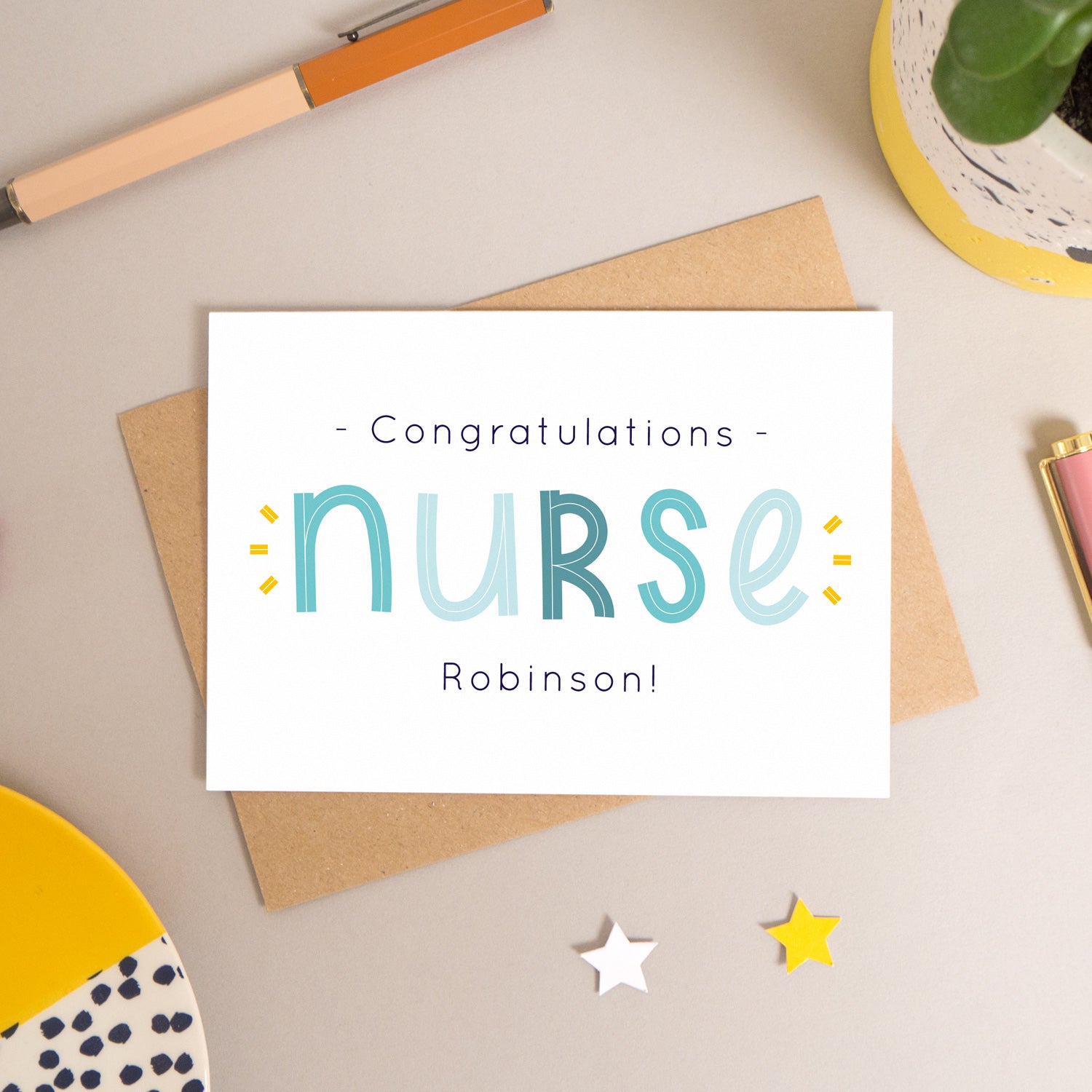 This personalised nurse card has been shot flatlay style looking directly over the top of the card. It is lying flat on it’s kraft brown envelope, on a warm grey background. Surrounding the card are pens, a clip, a plant and a trinket dish. This version of the card is in varying tones of navy and blue.