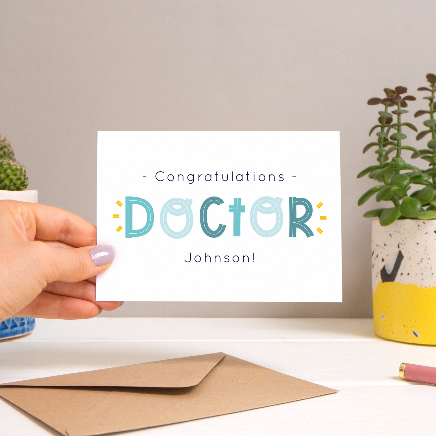 A personalised doctor card held over a warm grey and white background with potted plants peeping the sides. Behind the card is a kraft brown envelope that comes with the card. The text on this version of the card is in varying tones of navy and blue.